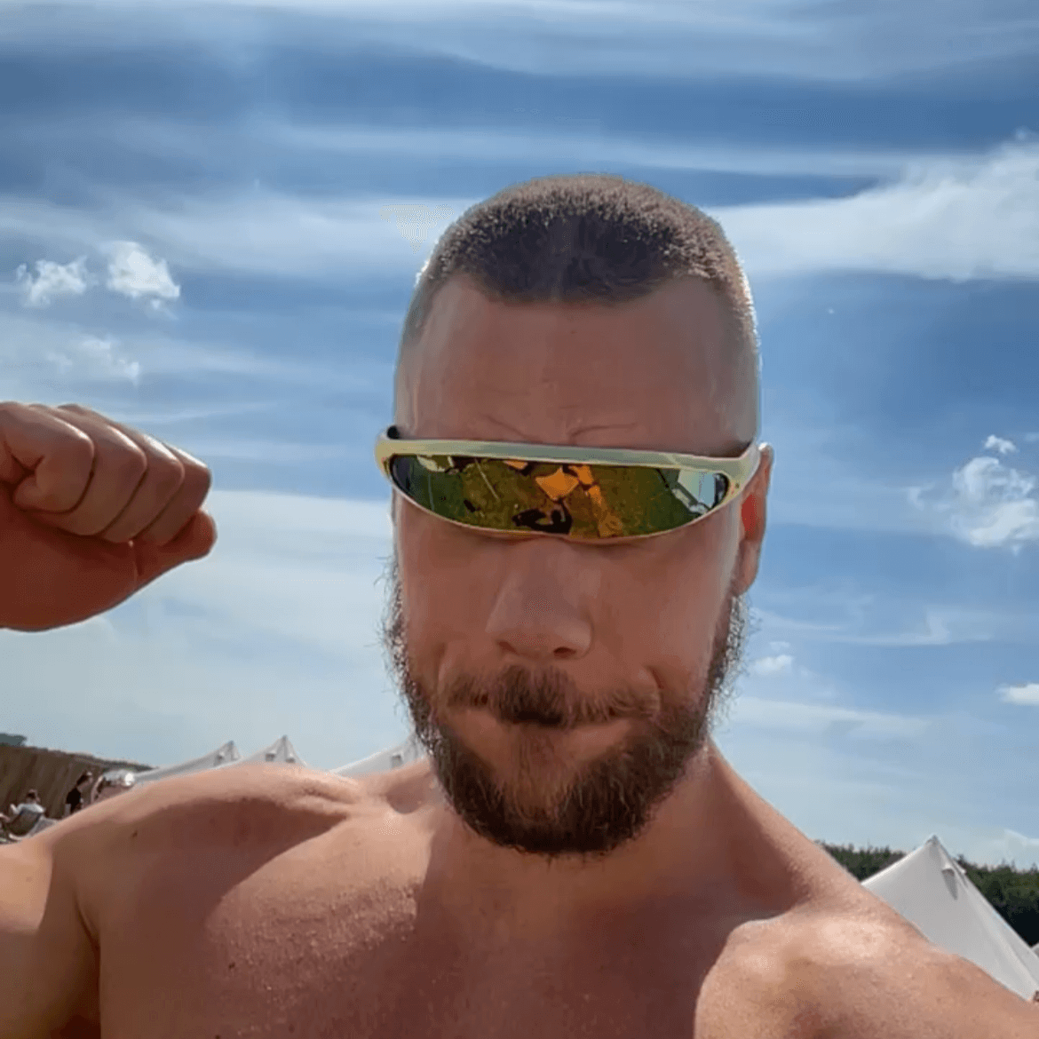 Man wearing futuristic RaveFather Cyber Matrix Cyclops sunglasses outdoors flexing arm under clear blue sky.