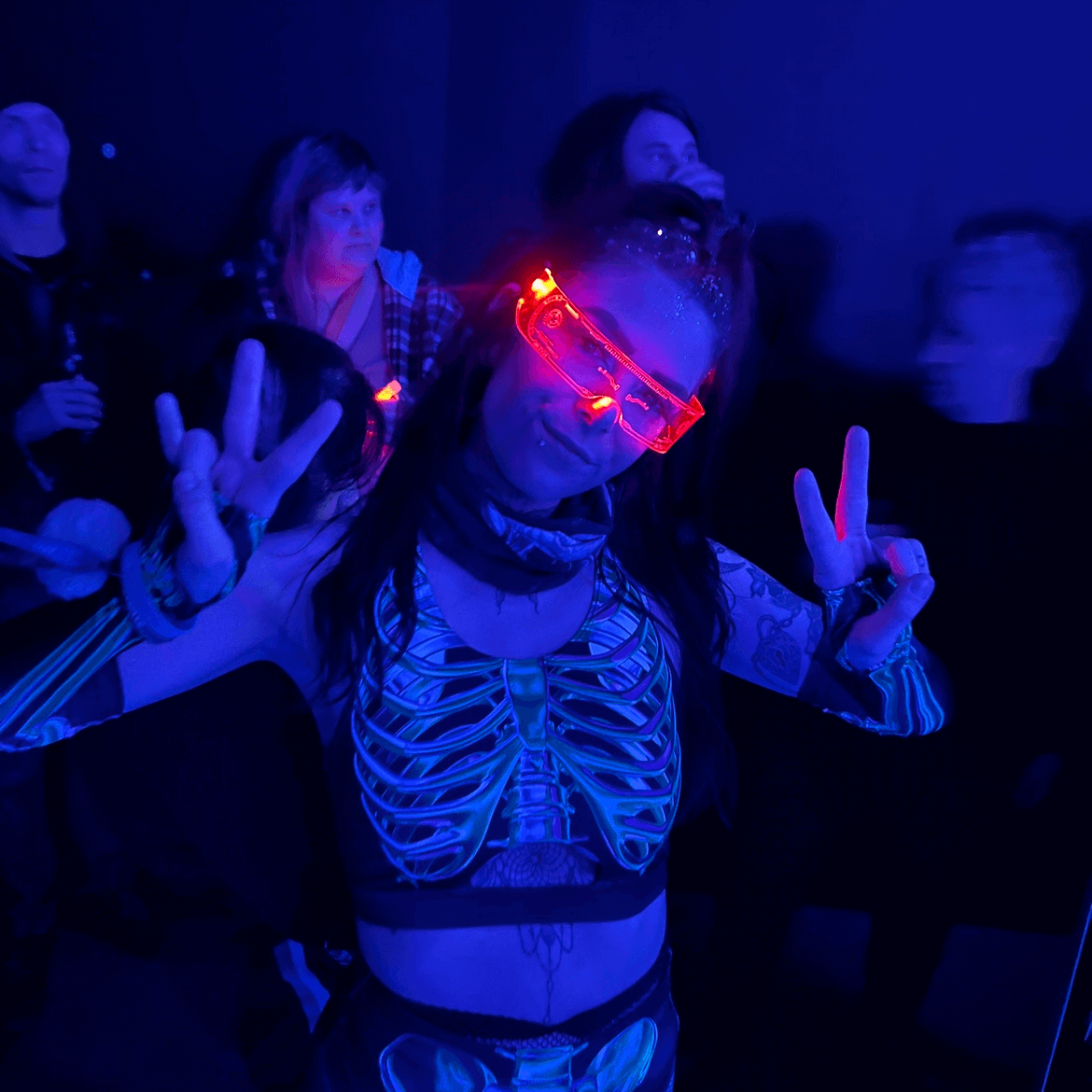 Person wearing RaveFather SpectrumShift LED Rave Vision glasses displaying peace signs at a rave party, glowing in vibrant light patterns.