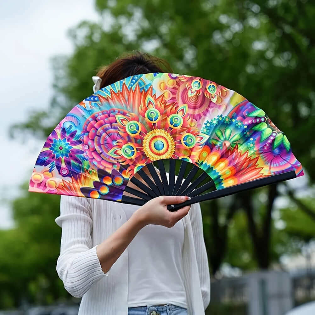 Person holding vibrant psychedelic UV glow folding fan at a festival, blending colorful designs with nature's green backdrop.